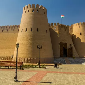 A high crenellated mudbrick fortress wall with tapering round towers and a carved wooden gate, a Tajik flag flying above and benches on the paving in front.