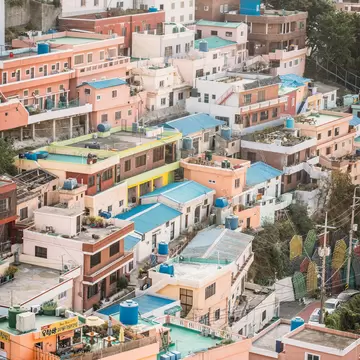Dense rows of pink, peach and white houses with blue roofs and water tanks climbing a steep hillside in Busan.