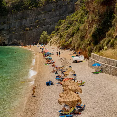 People under straw umbrellas on a sunny pebble beach.