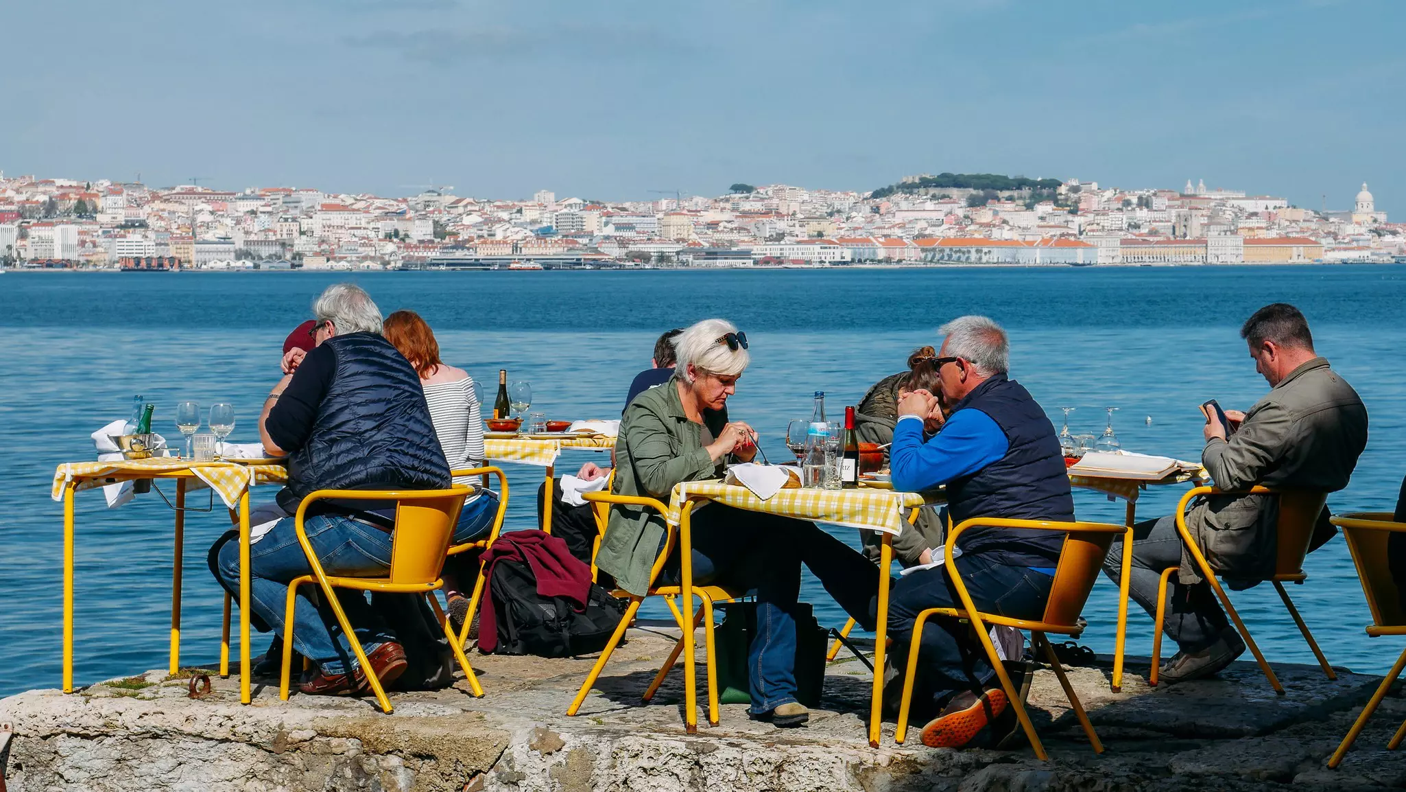 People sitting at yellow restaurant tables in the shore of the river Tagus in Cacilhas, with the cityscape of central Lisbon in the background