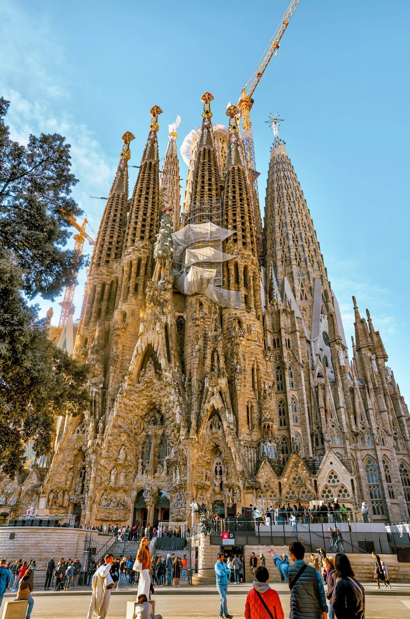 Tourists gather outside a cathedral with towering stone towers decorated with religious iconography and intricate sculptural details. Several cranes show the structure is having some building work.