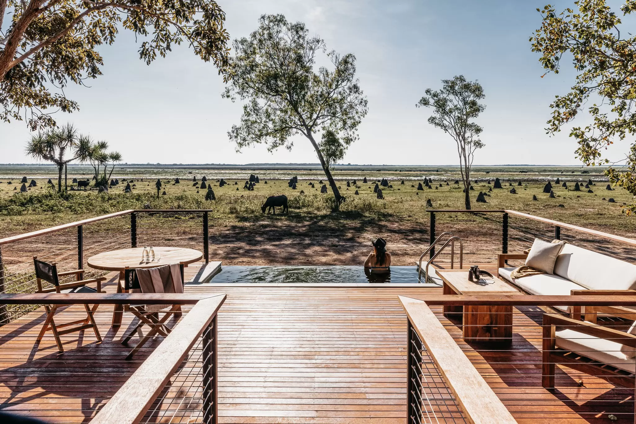 A wooden deck leads to a pool with a woman looking out at the landscape.