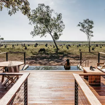 A wooden deck leads to a pool with a woman looking out at the landscape.