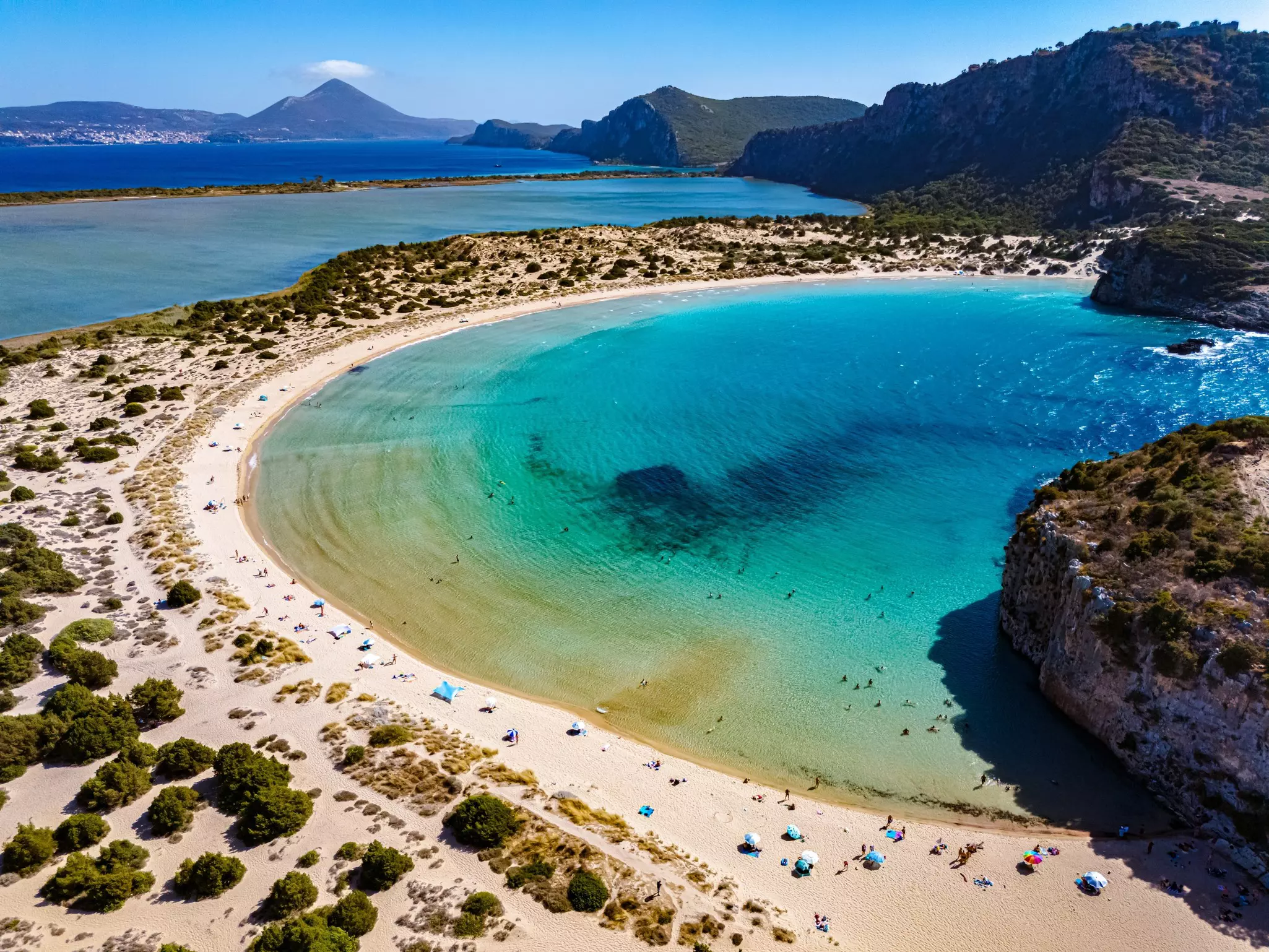 An overhead view of a curved beach enclosing a cove, with hills in the distance
