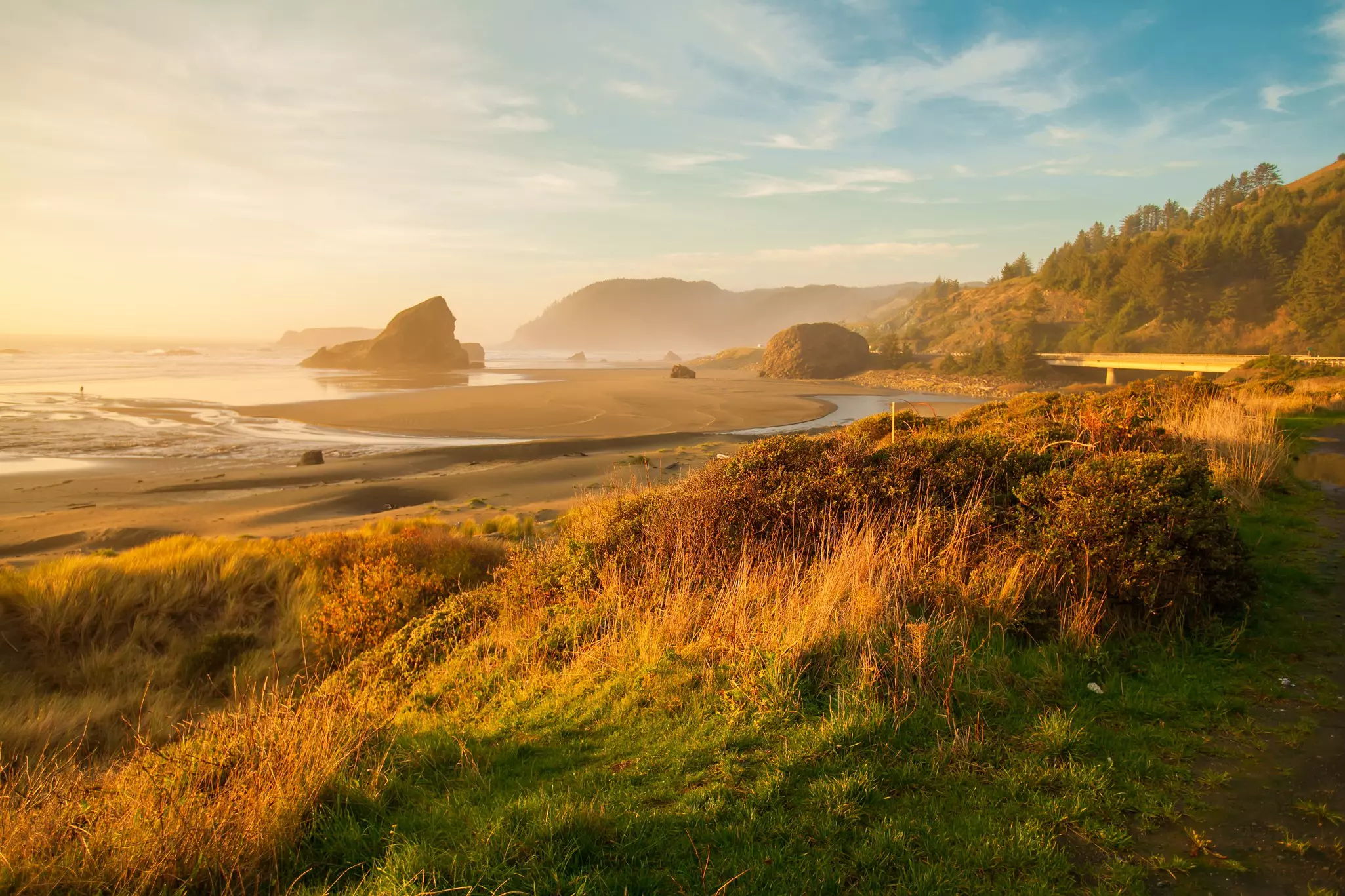 Grass-covered sand dunes give way to a vast sweep of sand with rocky outcrops at sunrise. A highway passes nearby.