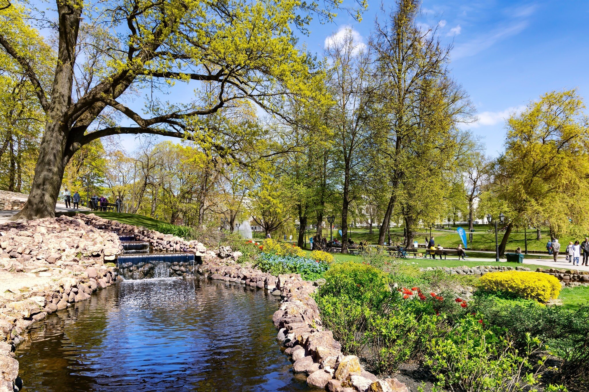 Small cascade flows into a stone-edged pond beneath budding trees, with tulips, benches and strolling people beyond