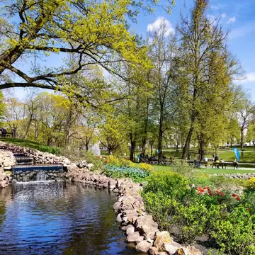 Small cascade flows into a stone-edged pond beneath budding trees, with tulips, benches and strolling people beyond