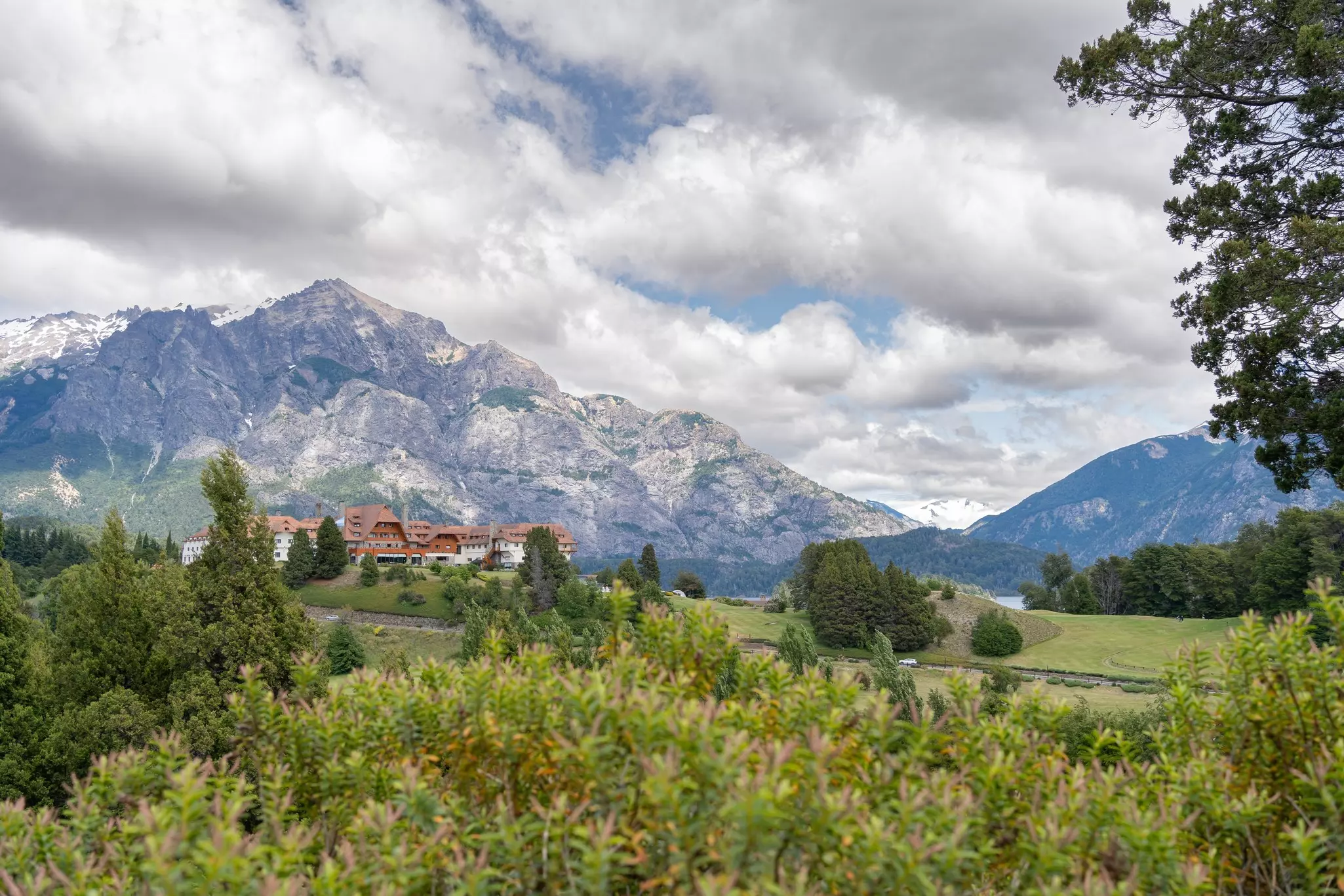 A large hotel at the base of a rocky mountain; a green field with bushes is in the foreground.