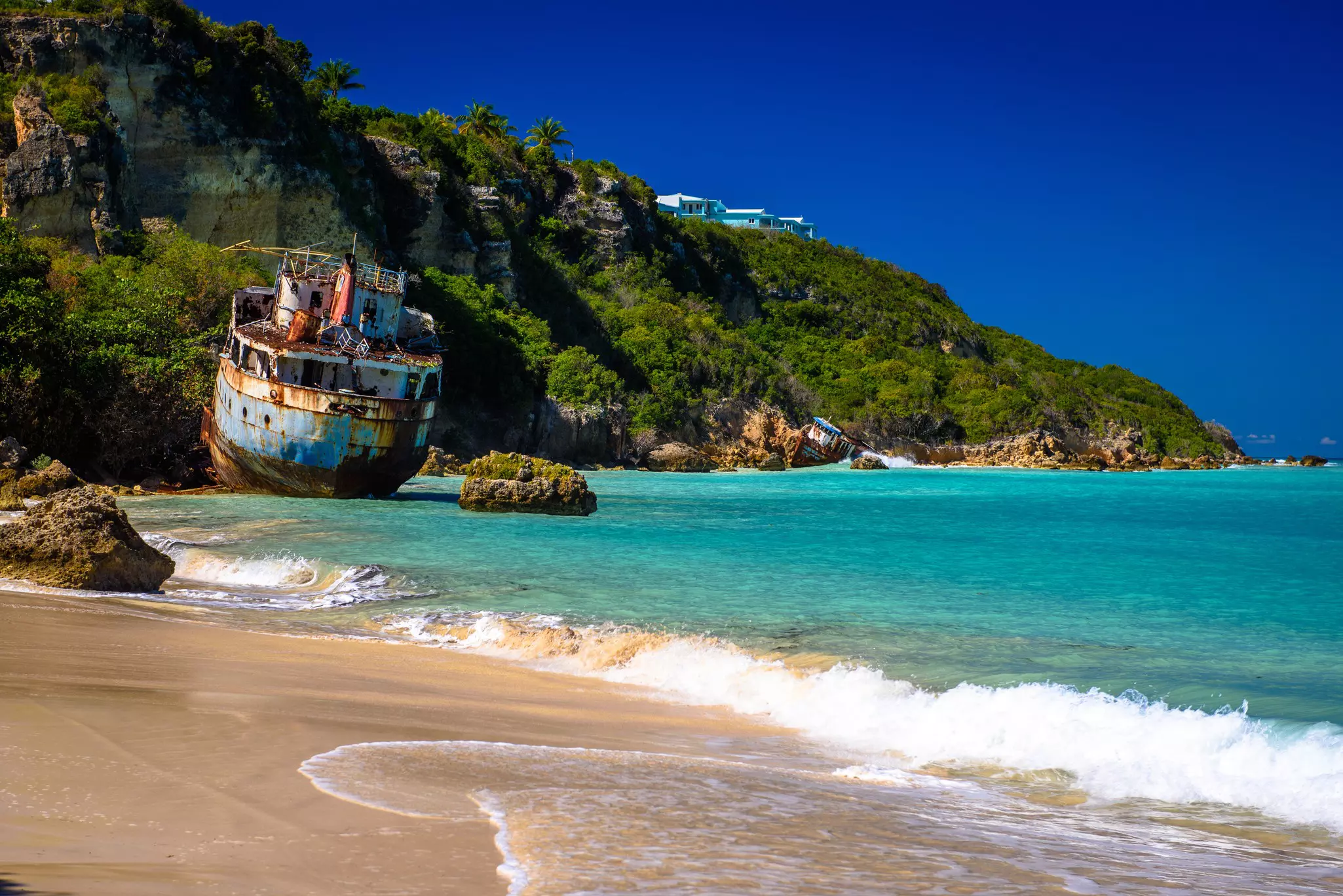 A large ship lies wrecked on the rocky shore near a sandy beach. Another smaller boat is on rocks further along the coastline.