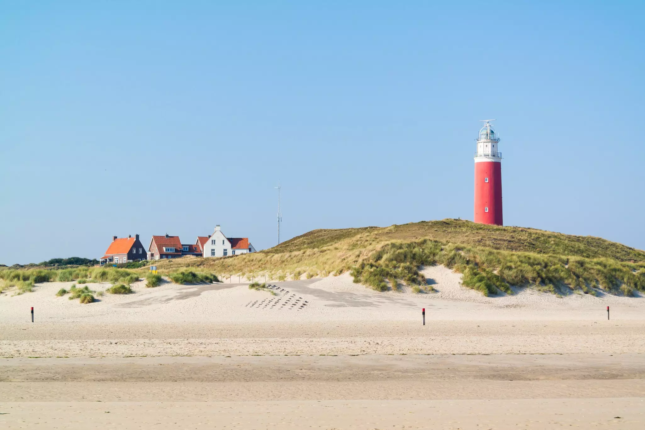 A sandy beach with a large grass-covered dune. Nearby stands a tall red lighthouse.