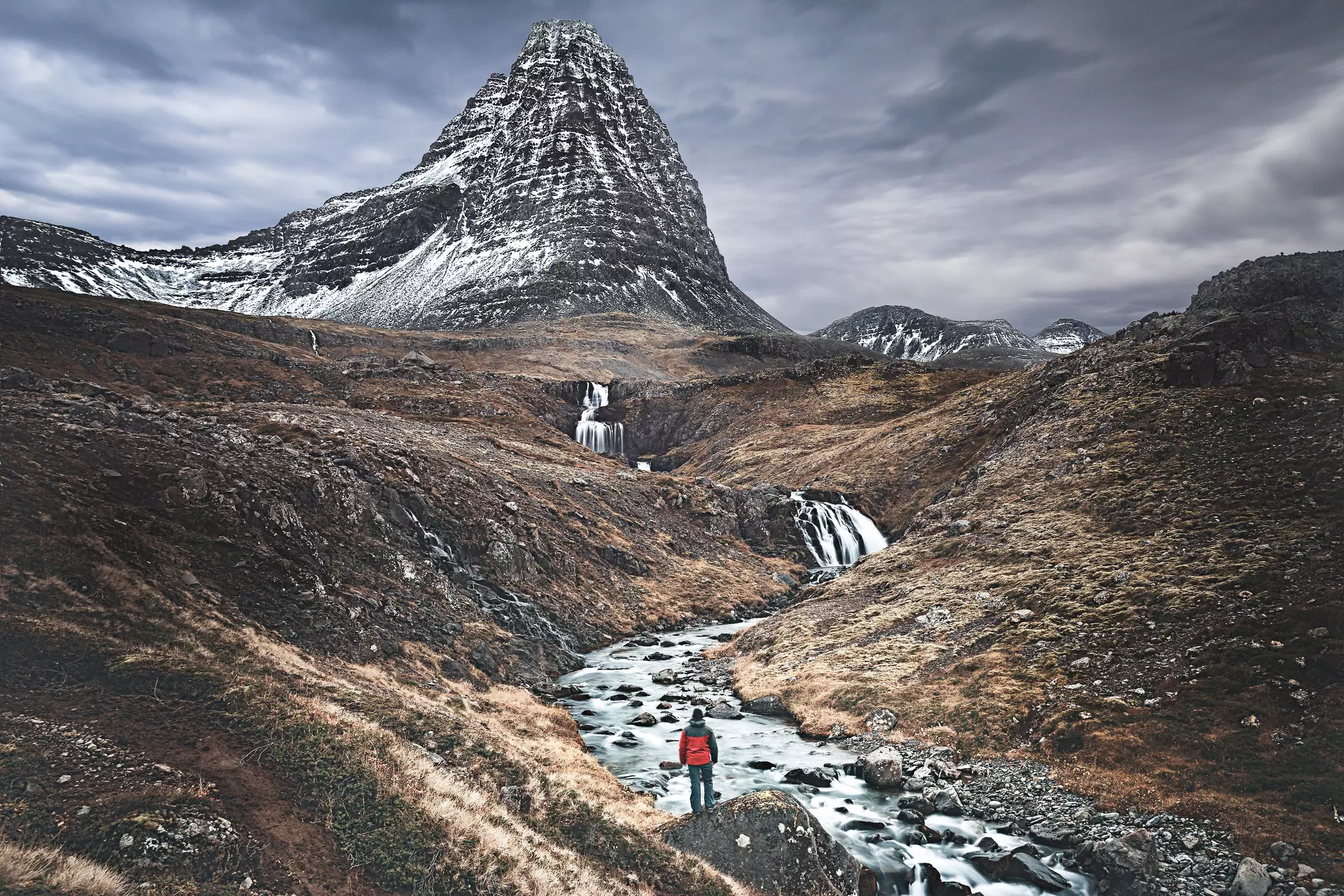 A lone hiker, seen from behind, stands on a rock overlooking a stream with waterfalls and snow-dusted mountains in the distance.