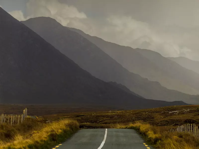 An empty road runs through a field heading towards hills and rain clouds
