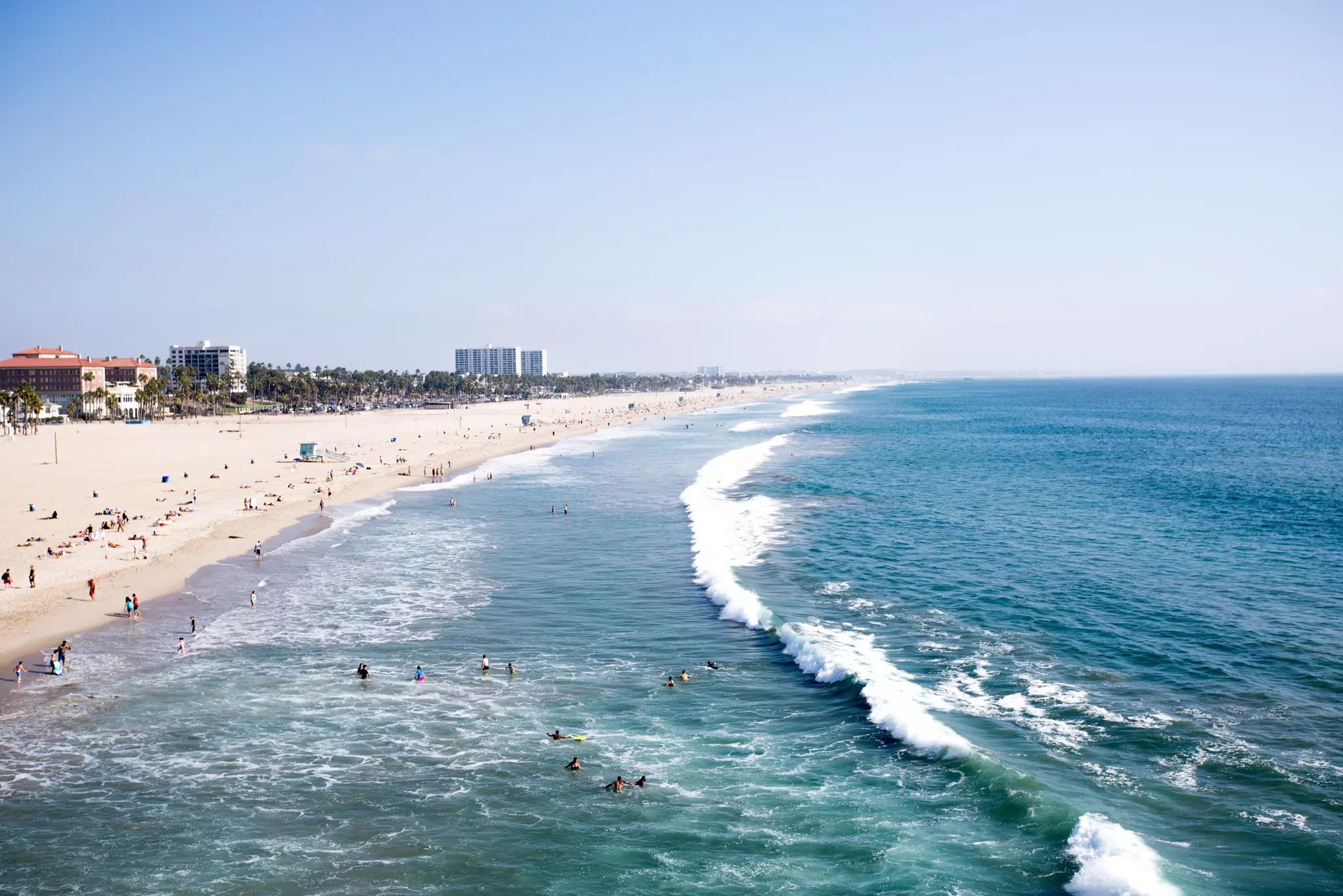 Surfers and swimmers in the ocean as large waves crash nearby. Sunbathers line the sandy shores on a vast beach dotted with lifeguard towers.