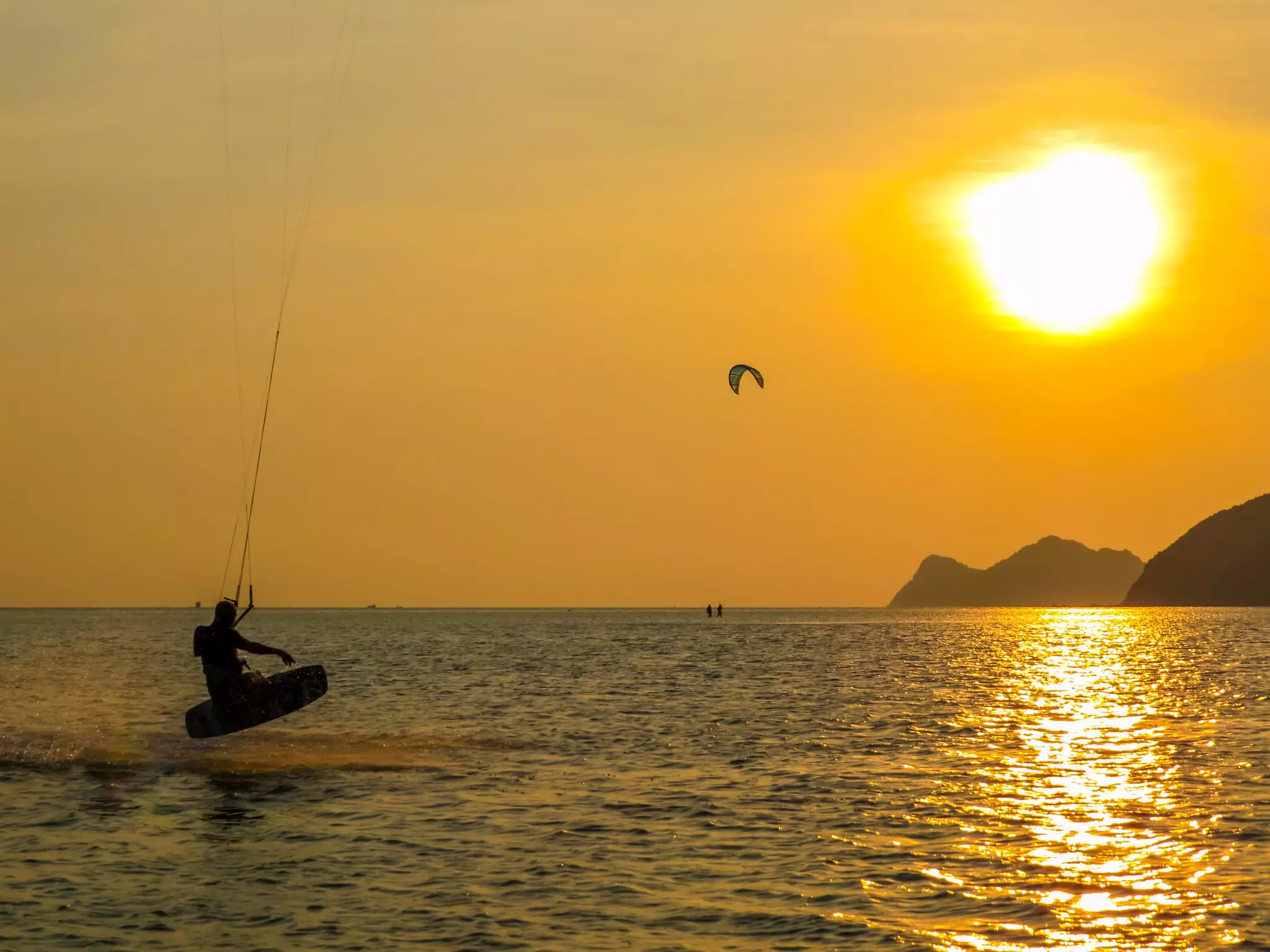 A kitesurfer leaps out of the water, silhouetted by the setting sun, whose rays turn the surface of the water gold.