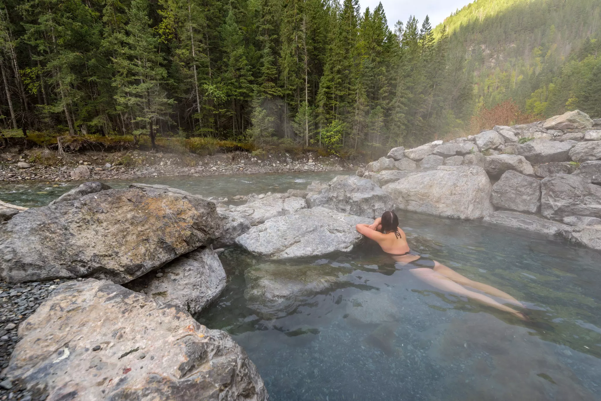 The back of a woman in a bikini stretched out in natural hot springs next to a river with evergreens in the distance.
