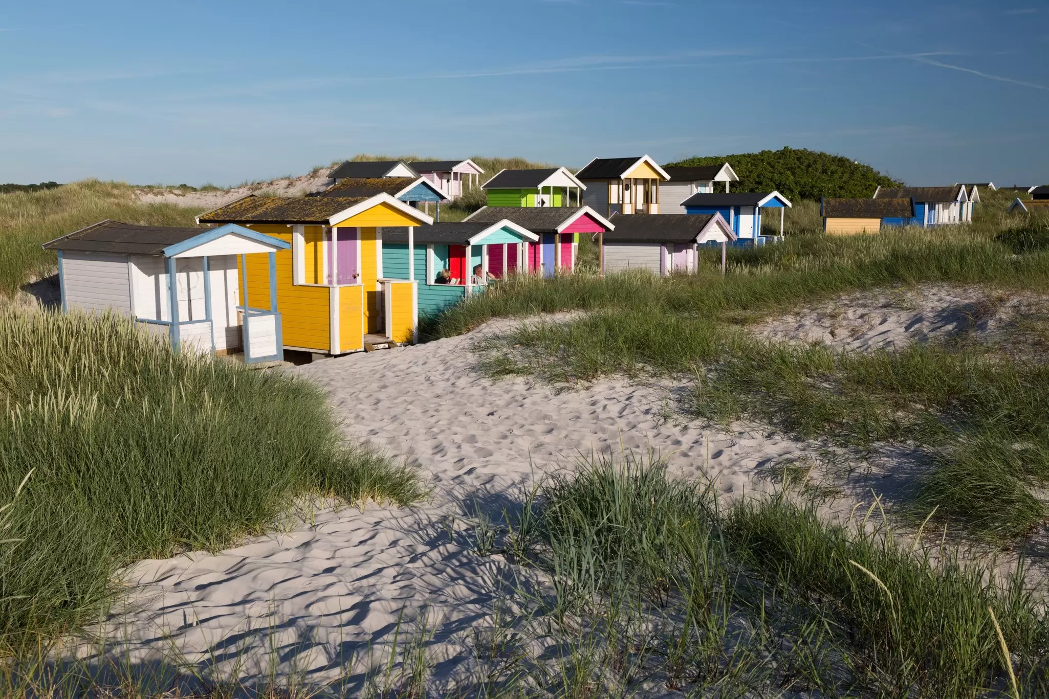 Colorful beach huts tucked into sandy and grassy dunes.