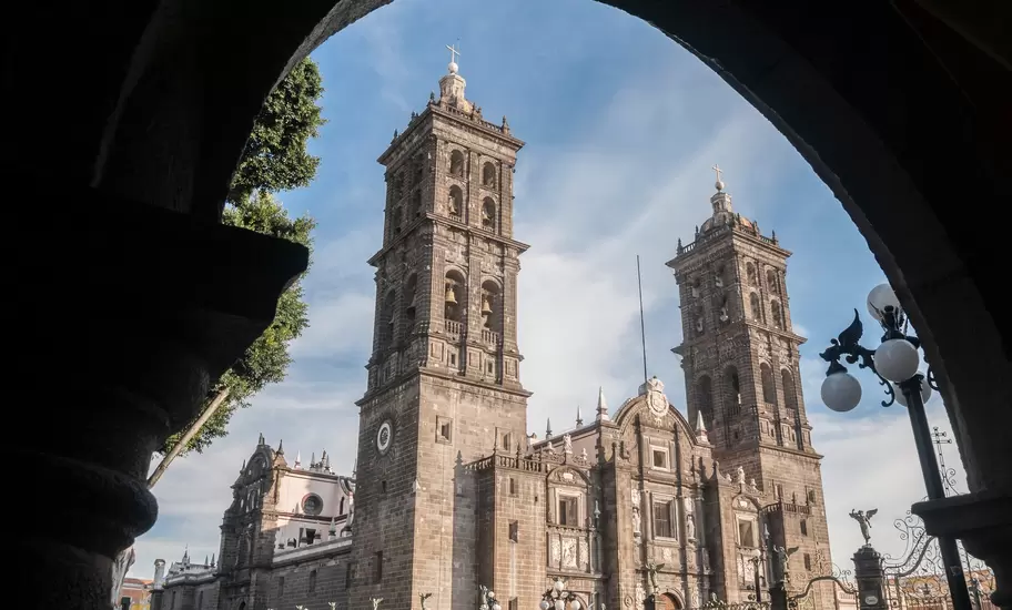 Puebla Cathedral's twin stone bell towers and facade, framed by a dark arcade arch, with an ornate iron lamp post