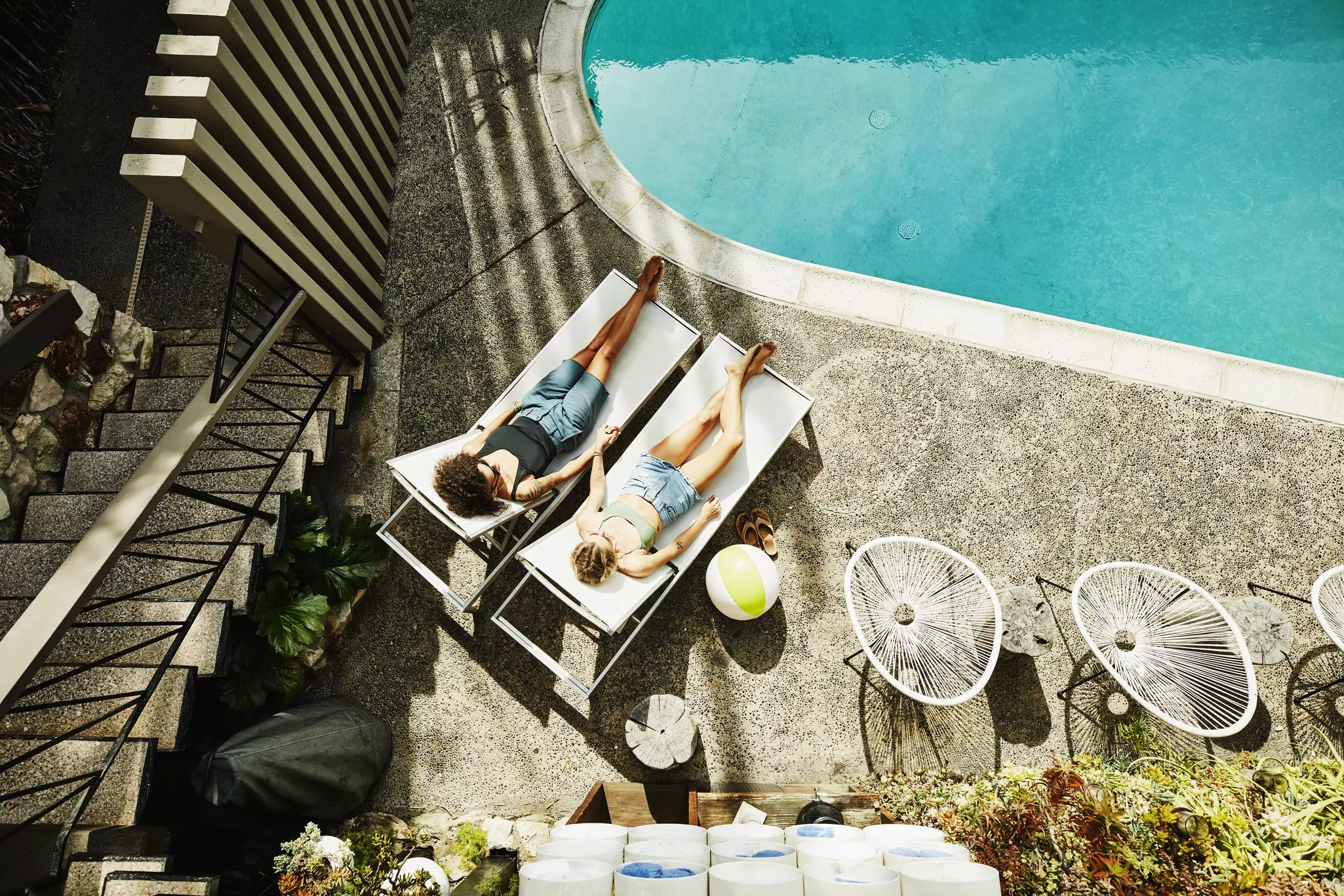 Overhead view of lesbian couple holding hands while relaxing in lounge chairs by hotel pool