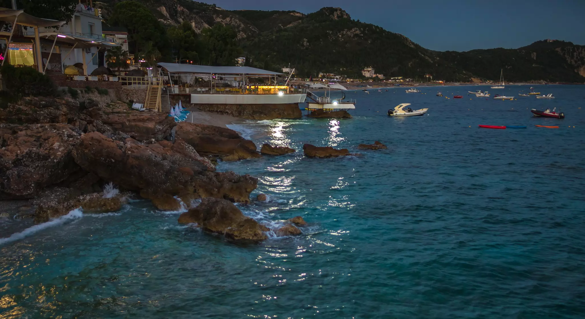 A rocky cove is pictured at night, with lights from a restaurant illuminating boats moored in the water.