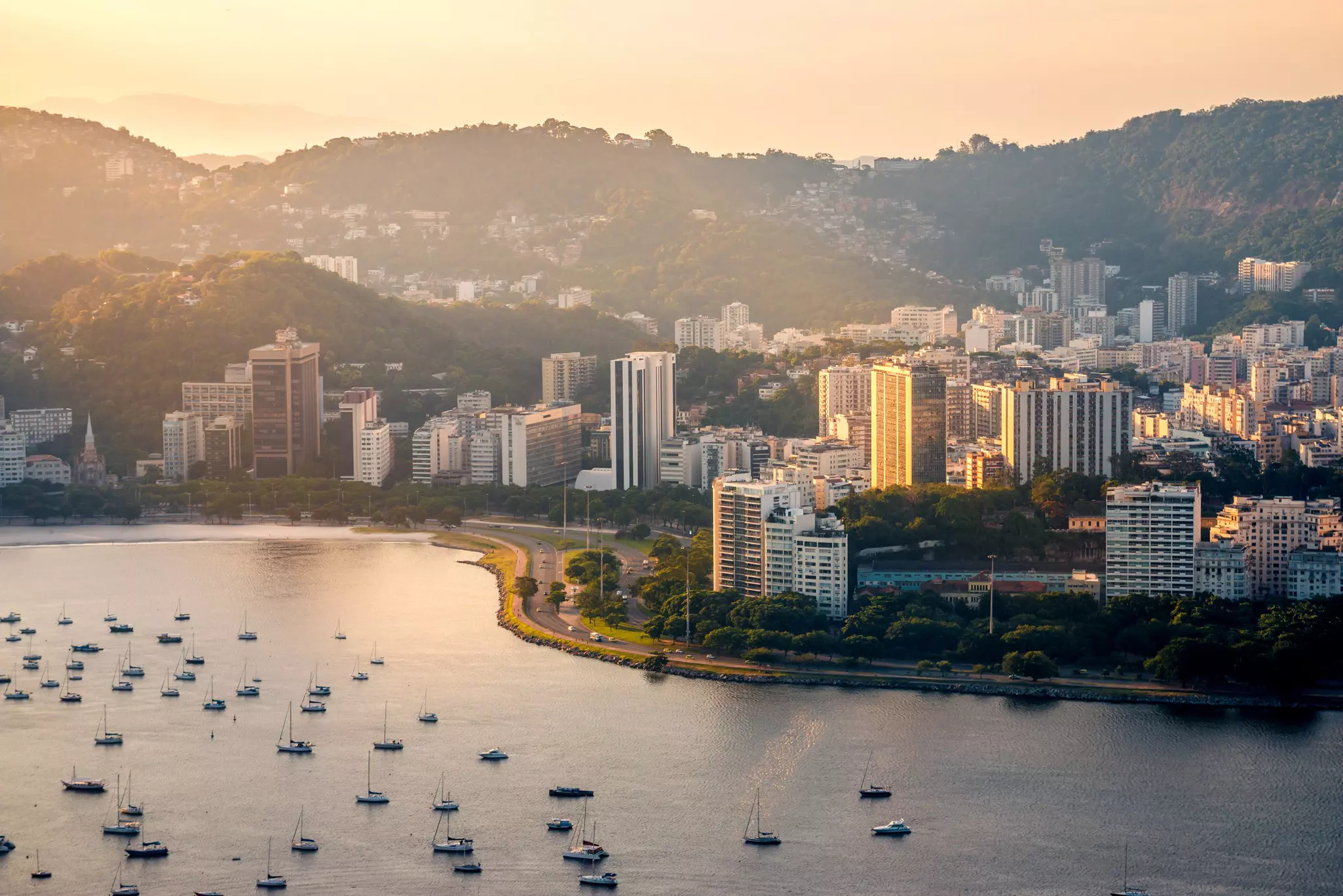 A view of a coast along a bay by dusk. Towers line the coast, boats are seen moored in the water, and buildings cover the hills of the city beyond.