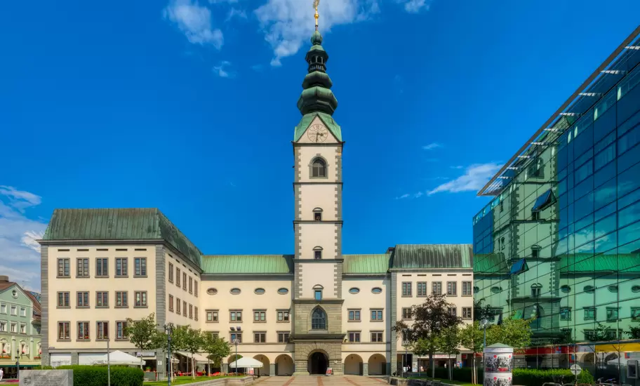 Klagenfurt Cathedral's clock tower with green onion spire on Domplatz, a glass office building reflecting it at right