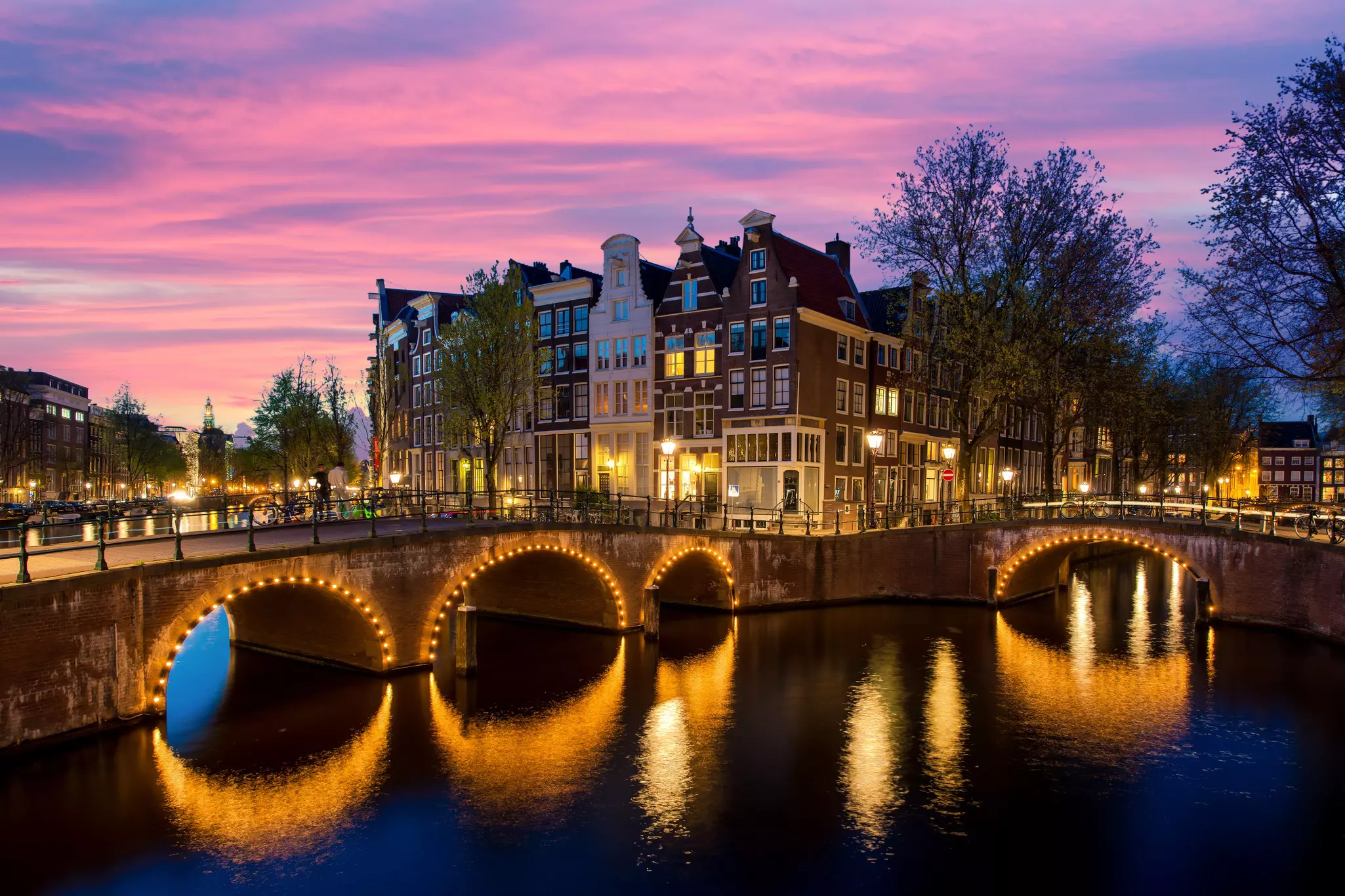 Low bridges at the intersection of two canals; the archways are lined with white lights, and the sky is a cloudy pink sunset.