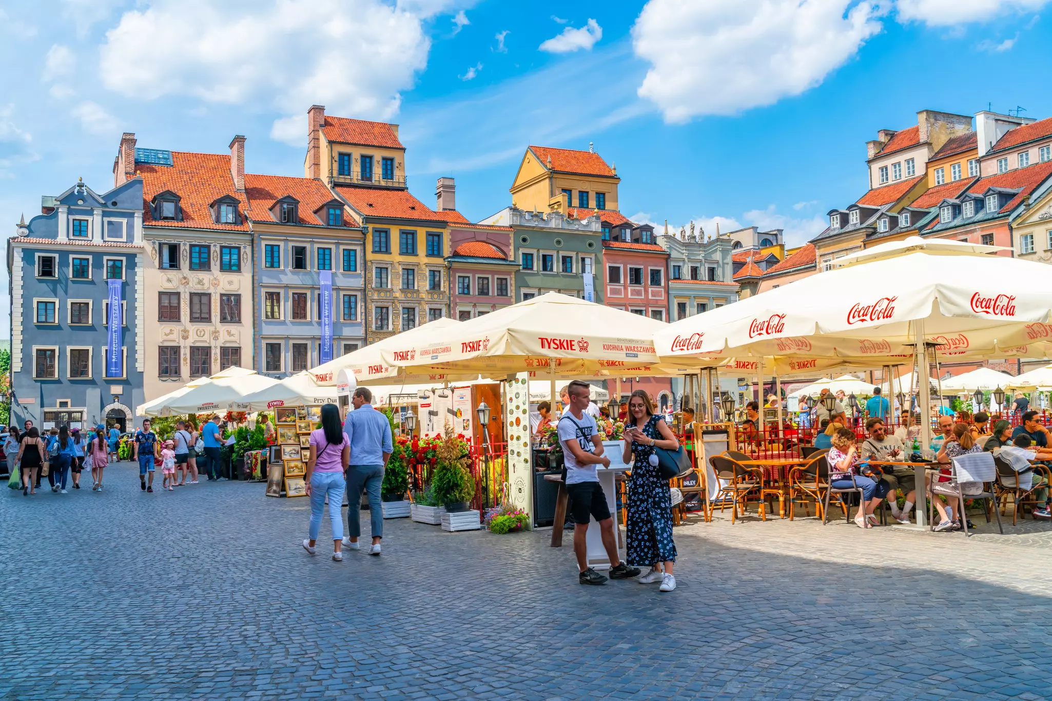 People walking around or sitting at tables under large umbrellas in a square surrouned by colorful buildings