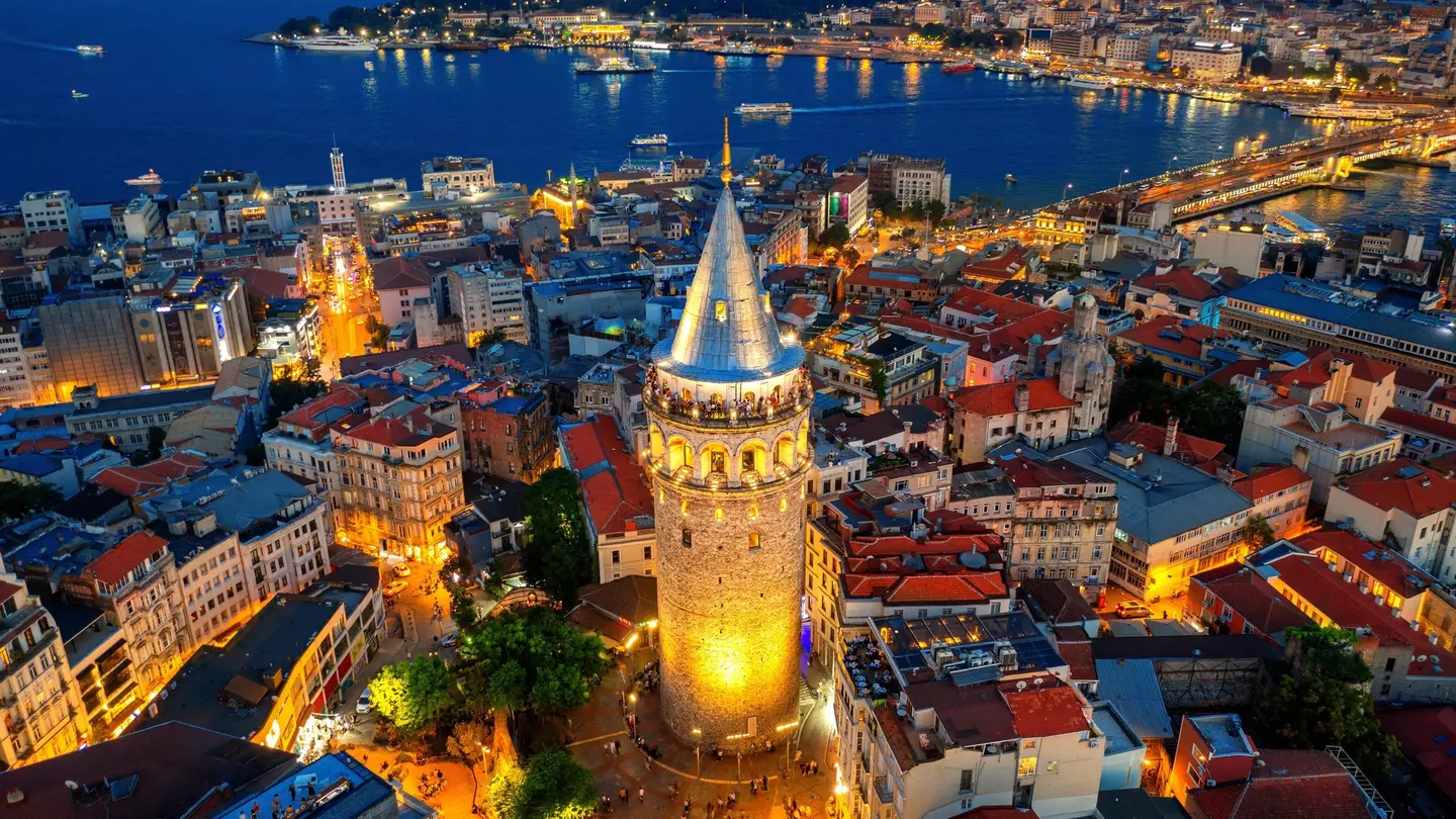An aerial view of a brick tower by a coastline in Türkiye at night; it is lit on its base and from within at the top. A city of low buildings surrounds the tower.