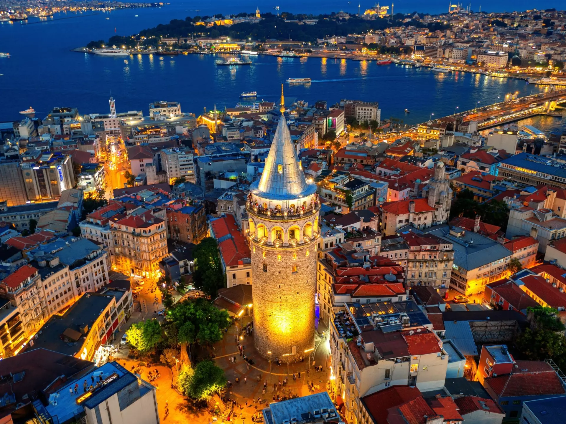An aerial view of a brick tower by a coastline in Türkiye at night; it is lit on its base and from within at the top. A city of low buildings surrounds the tower.