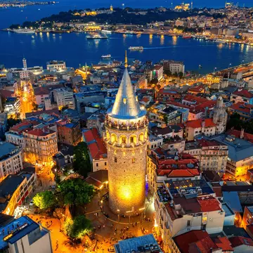 An aerial view of a brick tower by a coastline in Türkiye at night; it is lit on its base and from within at the top. A city of low buildings surrounds the tower.