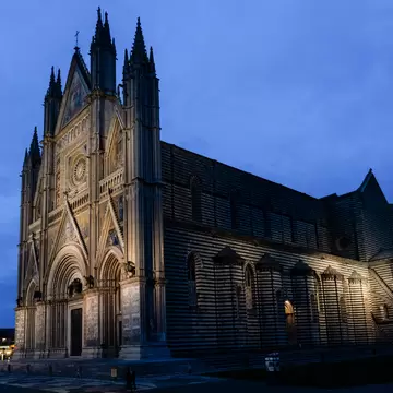 Orvieto's Gothic cathedral under a deep blue evening sky, its spired front and striped side wall lit against the dark.