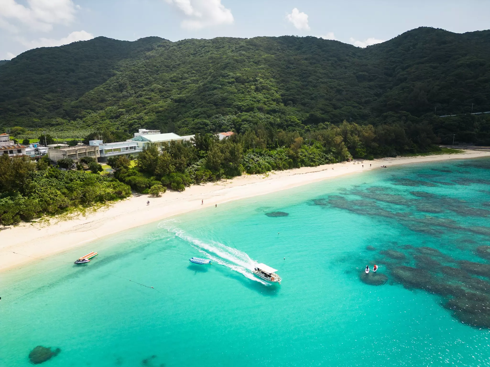A ferry boat in turquoise water leaving a white sand beach; a few buildings are in lush greenery off the beach.