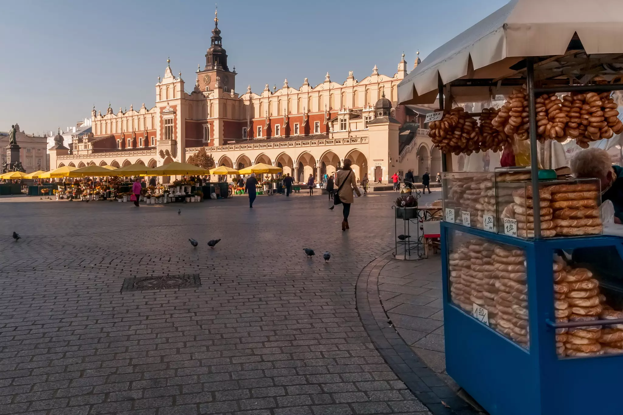 A vendor selling bread from a blue cart at the edge of a large square; yellow umbrellas at a cafe in the square and a long building with archways are in the background.