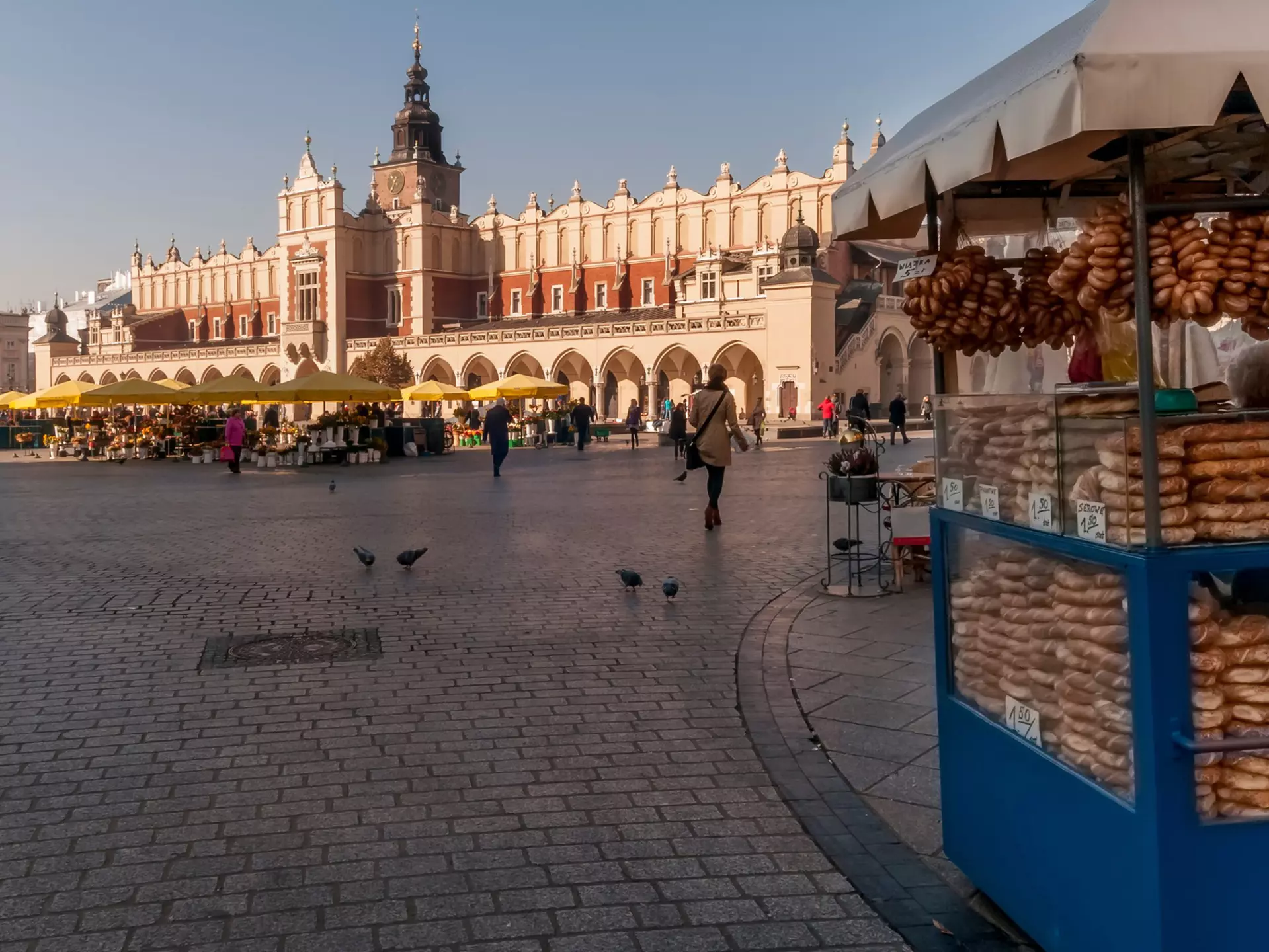 A vendor selling bread from a blue cart at the edge of a large square; yellow umbrellas at a cafe in the square and a long building with archways are in the background.