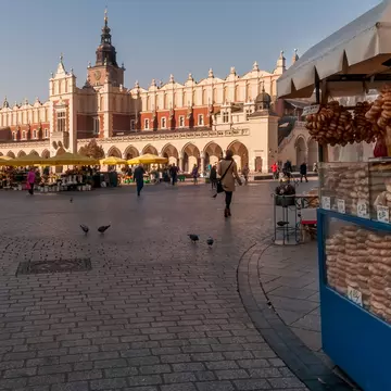 A vendor selling bread from a blue cart at the edge of a large square; yellow umbrellas at a cafe in the square and a long building with archways are in the background.