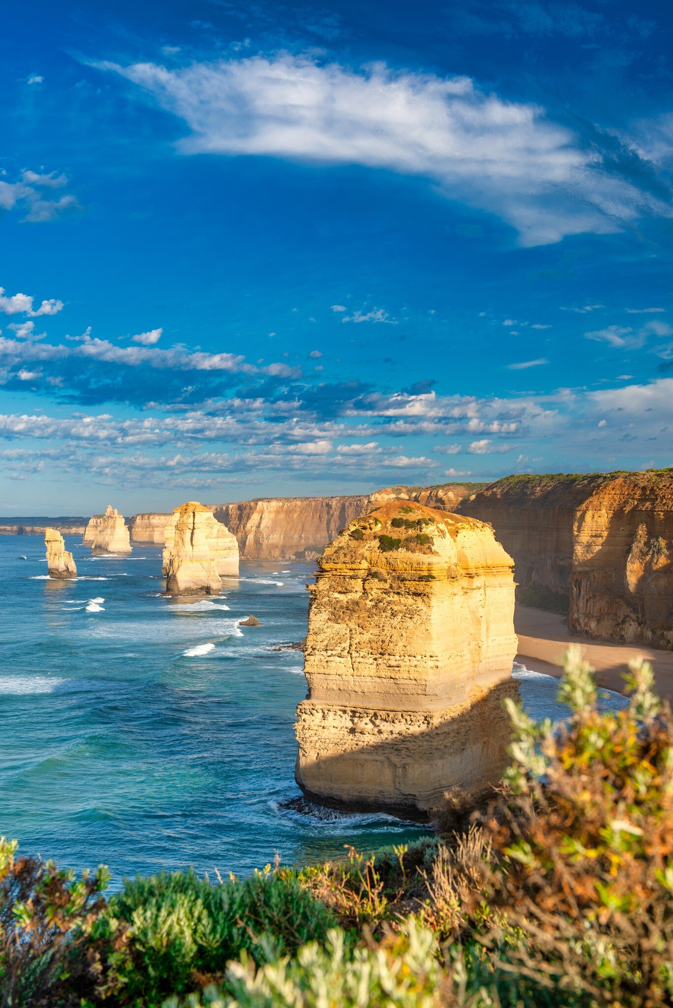 Sunlit rock stack in blue-green surf beside tall cliffs, with more stacks along the coast under a cloud-streaked sky