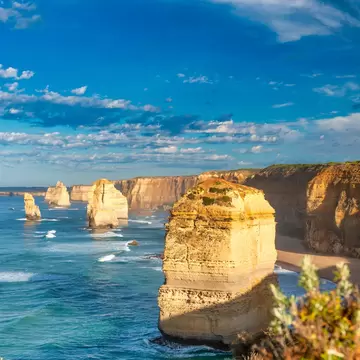 Sunlit rock stack in blue-green surf beside tall cliffs, with more stacks along the coast under a cloud-streaked sky