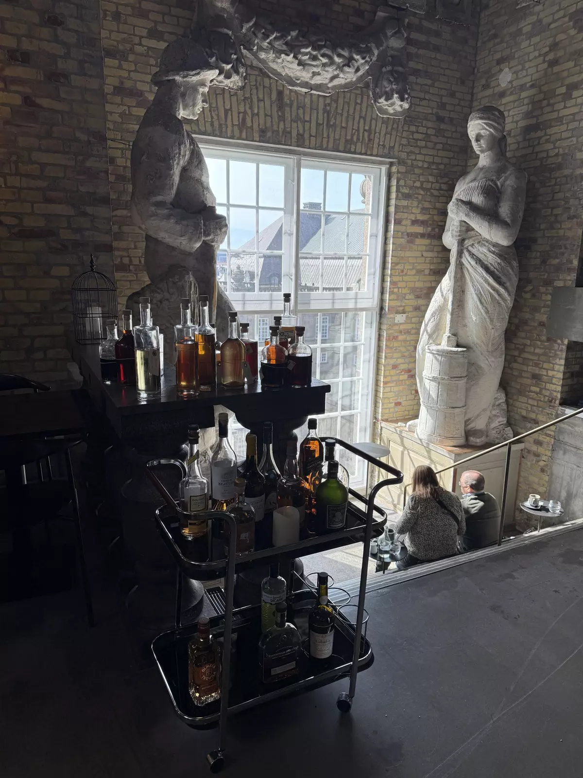 Close-up of bar and rolling tray containing bottles of alcohol with tall, white sculptures of people surrounding a tall window and a couple sitting on cement stairs.Inside the tower at Tårnet in Cophenhagen