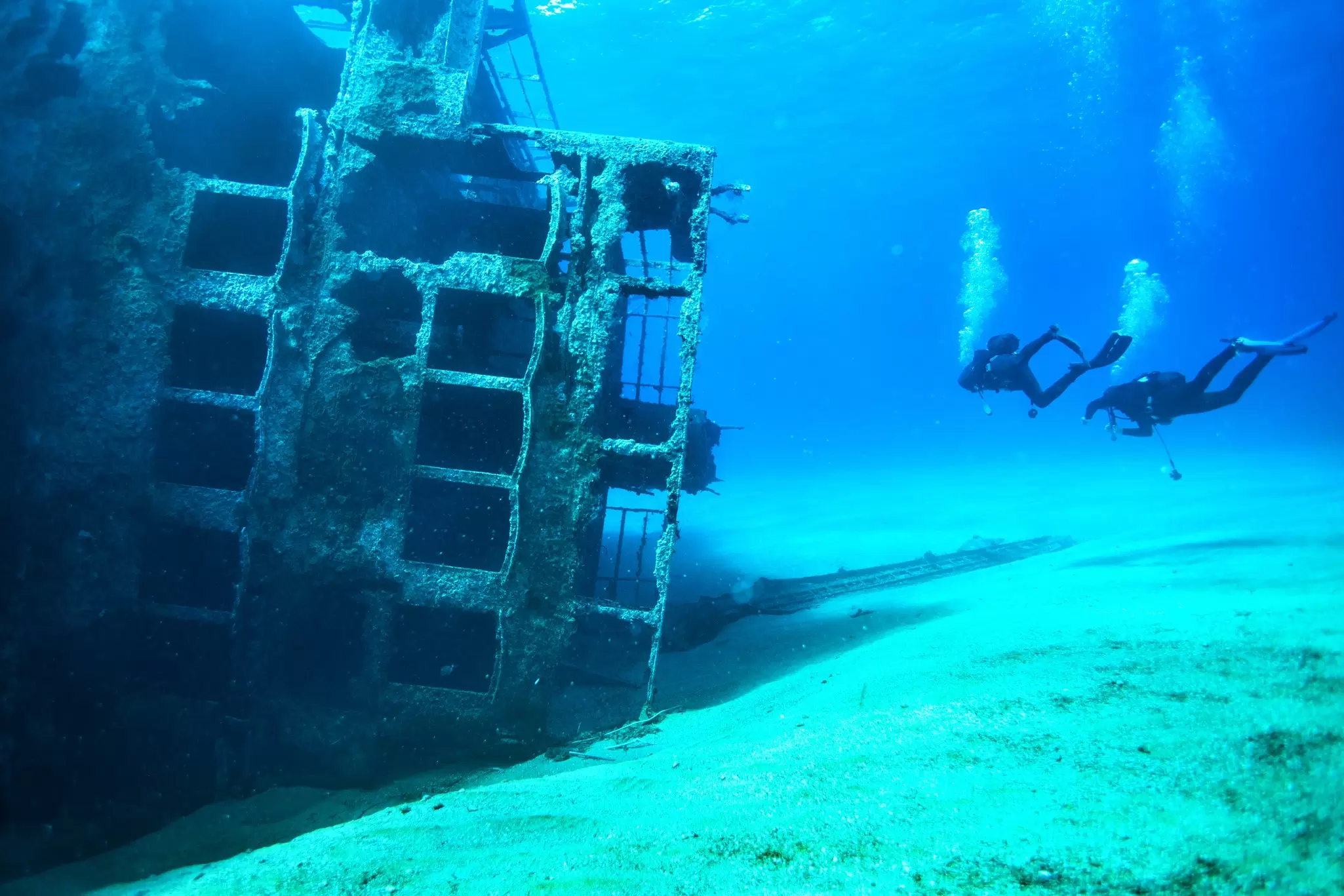 Two scuba divers descend towards a shipwreck lying on its side on the sandy seabed in Greece