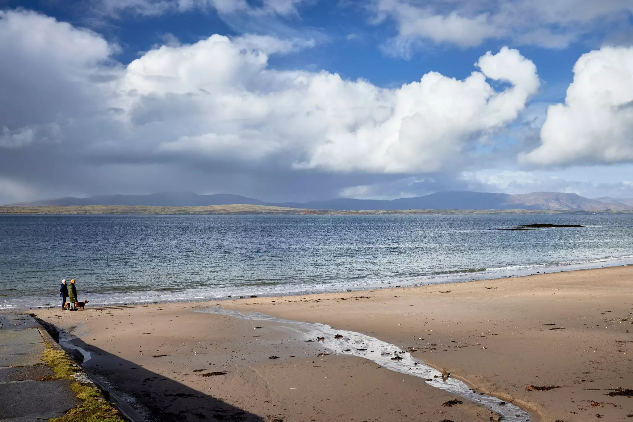 People walking a dog on a sunny day at Ganavan Sands, Oban, Scotland.