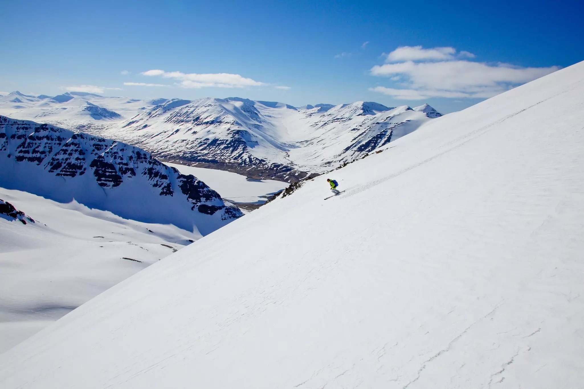 A lone skier is seen in the distance on a steep slope with snowy mountains beyond.