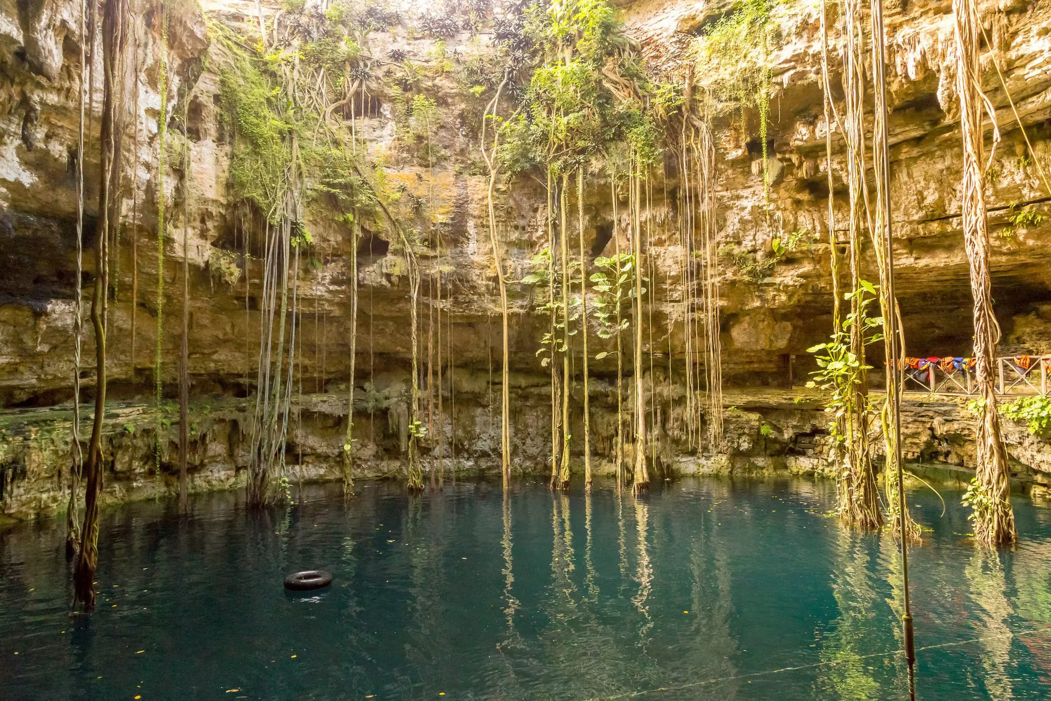 The deep blue waters of the cenote at Hacienda San Lorenzo are surrounded by buff and off-white stone walls strewn with long vines dangling from the top of the frame to the surface of the water. To the left, a black inner tube floats on the water.