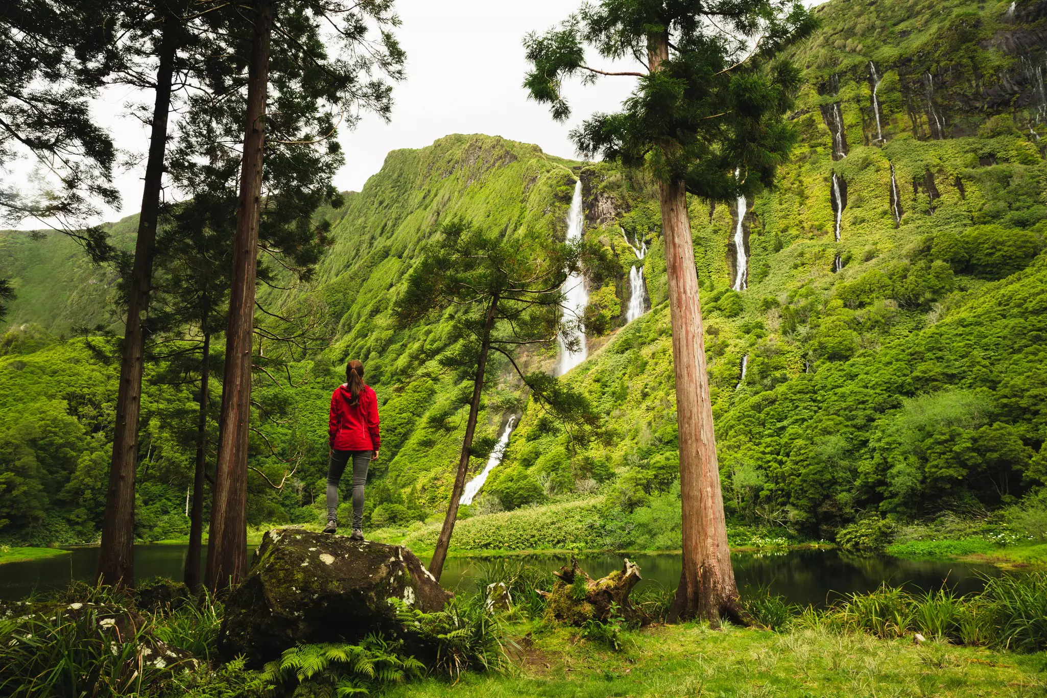 A woman stands on a rock and looks at waterfalls among a forest and green-covered mountains.