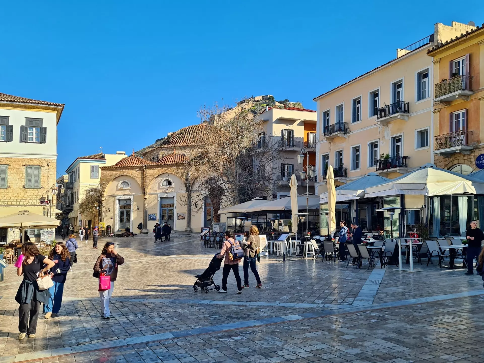 People walk through the square of a city in winter. A fortress on a hilltop is seen in the distance.