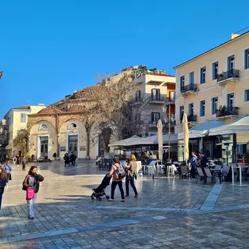 People walk through the square of a city in winter. A fortress on a hilltop is seen in the distance.