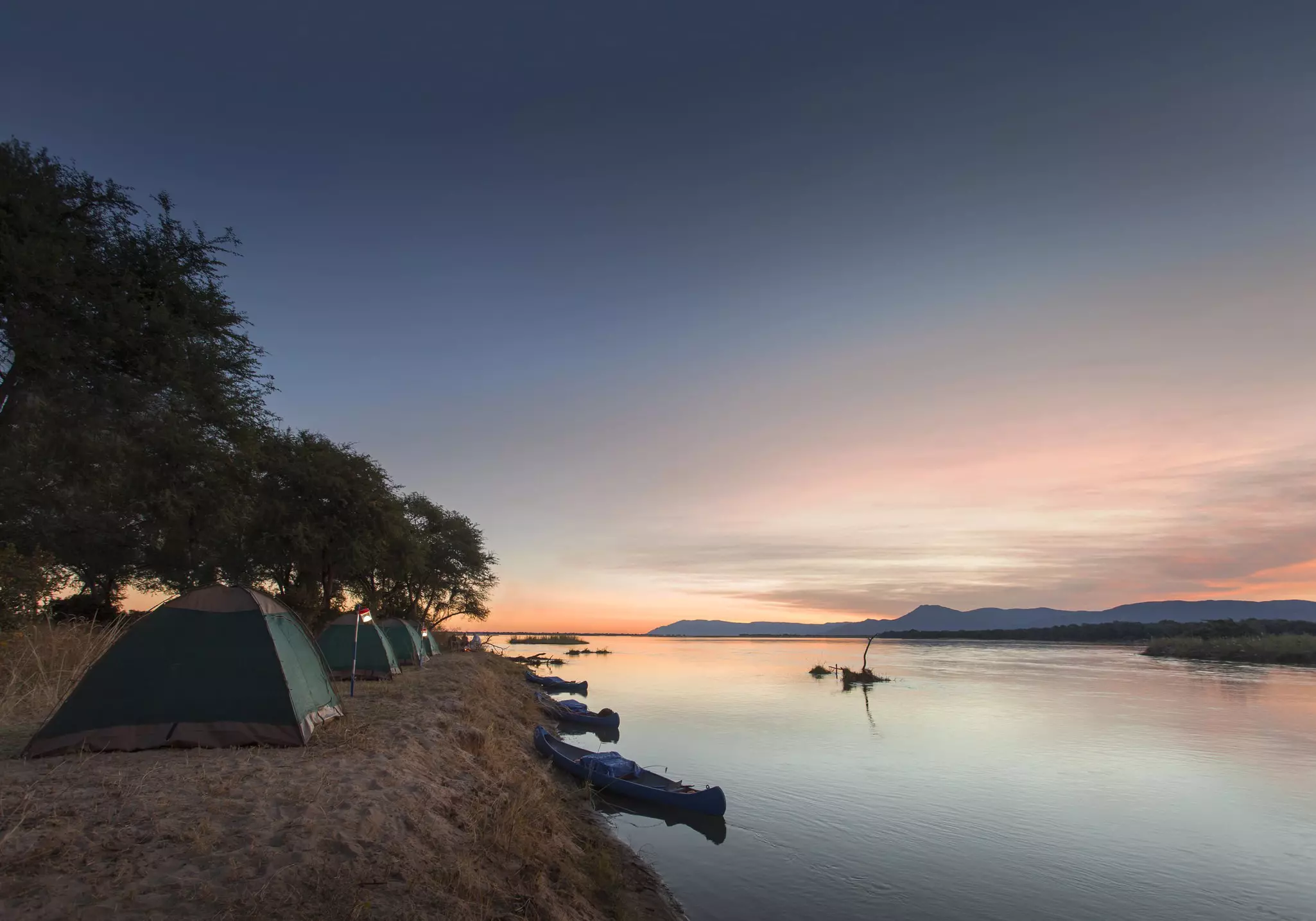 A group of tents sits in a line along the edge of the Zambezi river at sunset; trees flank the tents and canoes are moored on the steep bank.