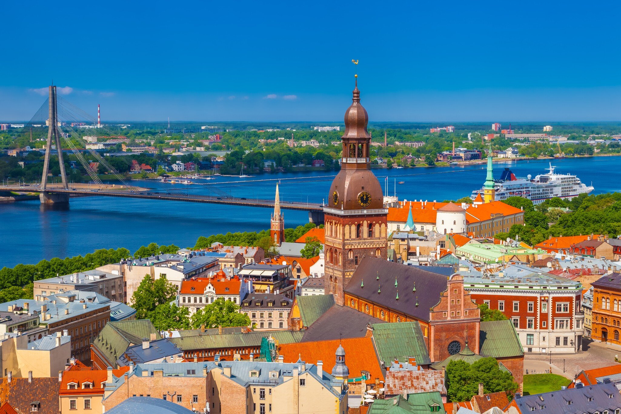 Brick cathedral with a green-brown domed tower among colourful rooftops, a wide river and cable bridge behind.