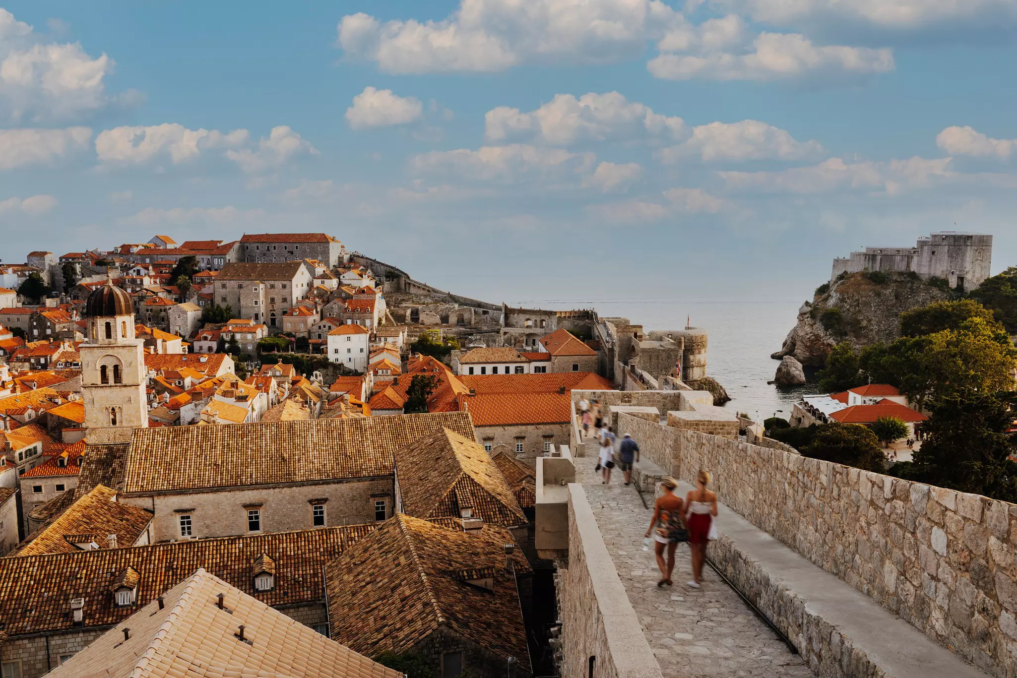 People move along a stone walkway that runs along the edge of the city walls that encircle a historic red-roofed settlement by the sea.