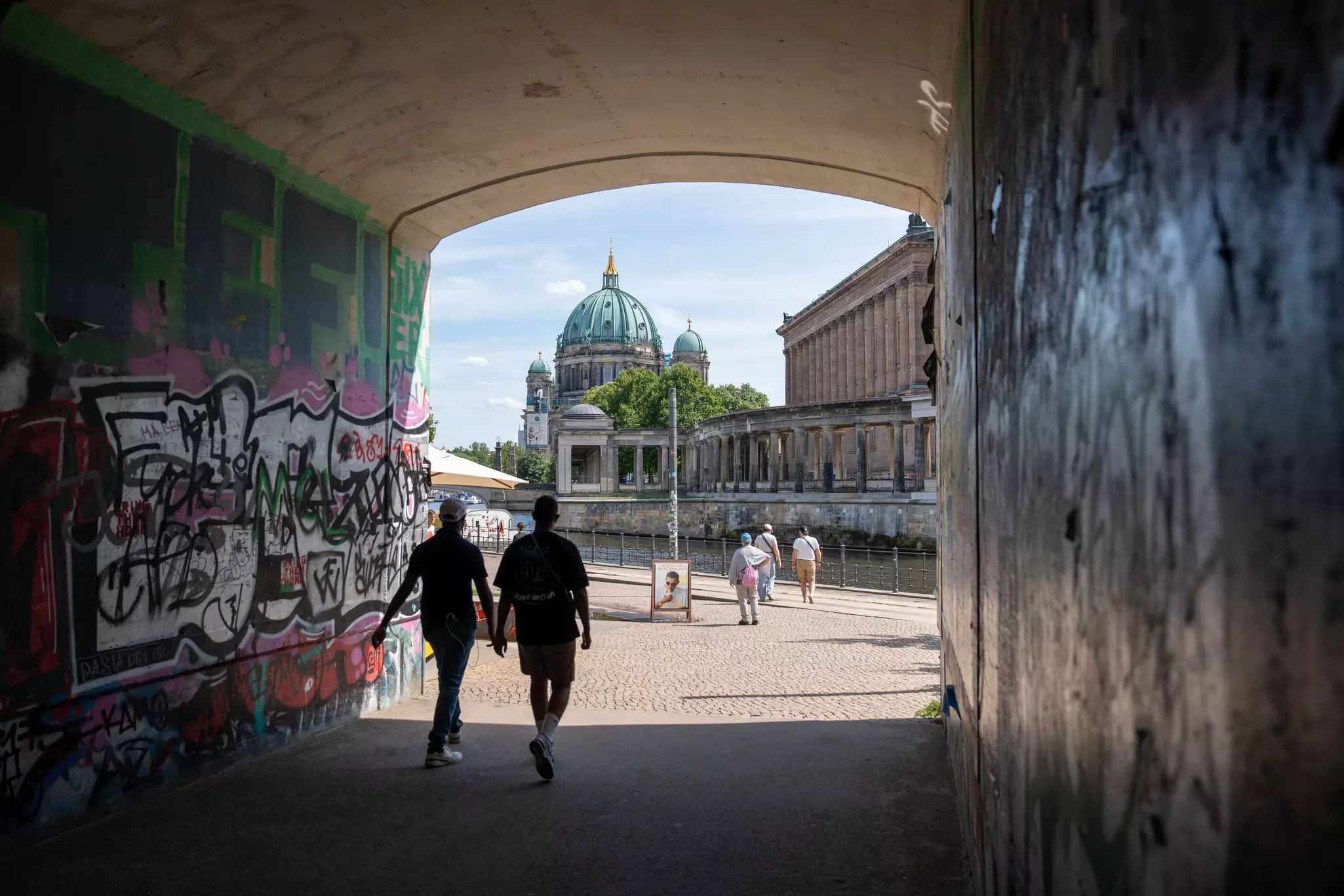 People walk through a passageway toward a narrow river and a green-domed building in a city.