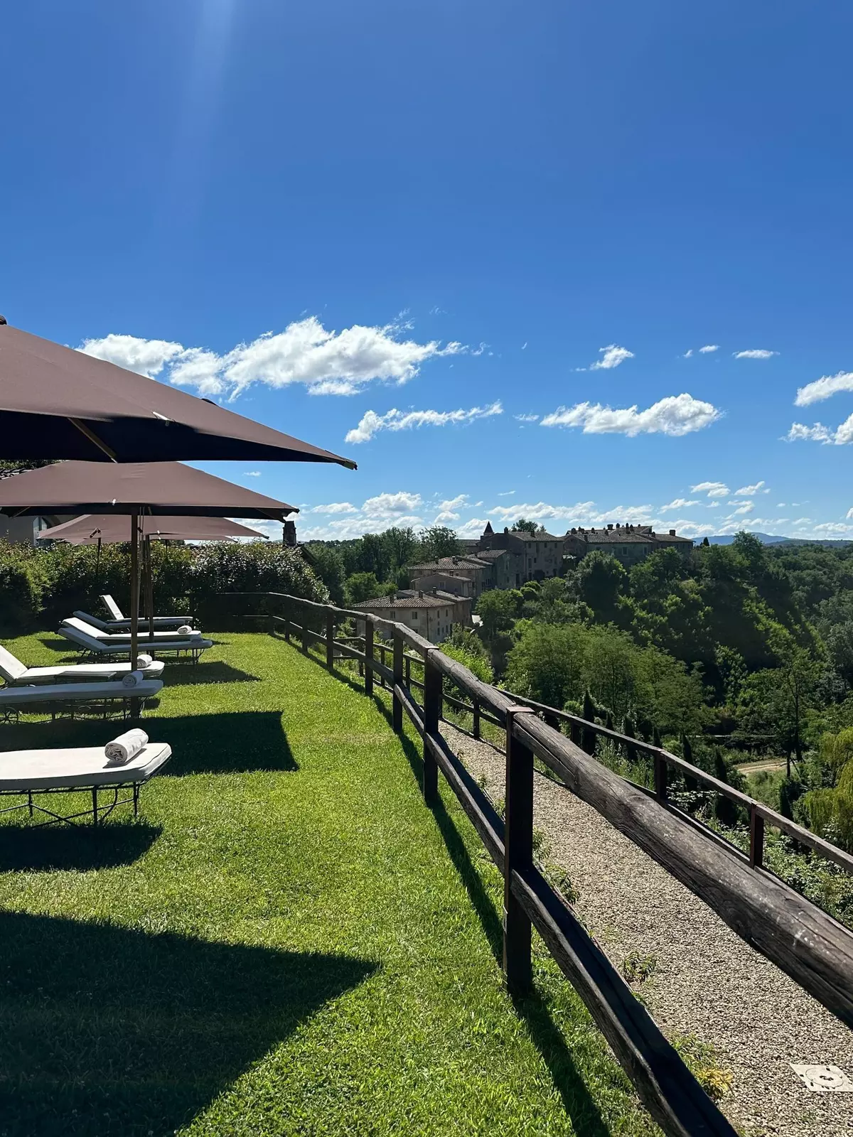 Lounger chairs in grass with umbrellas face a forested area; stone buildings are in the distance.